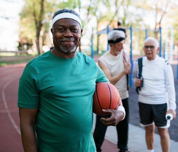 Portrait of a cheerful senior man exercising outdoors with his senior male friends, leading a healthy lifestyle and holding a basketball, smiling