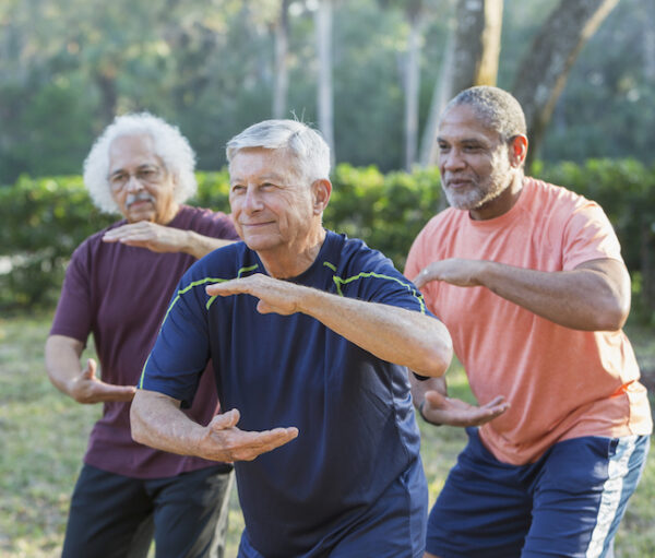 A group of three multi-ethnic senior men in the park practicing tai chi. The focus is on the caucasian man standing in the middle. He is in his 70s.