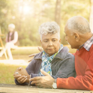 Mixed race senior adult couple talk at outdoor park in spring or autumn season. They sit on a park bench and discuss their relationship difficulties. African and middle eastern descent couple. Woman painting using easel in background.